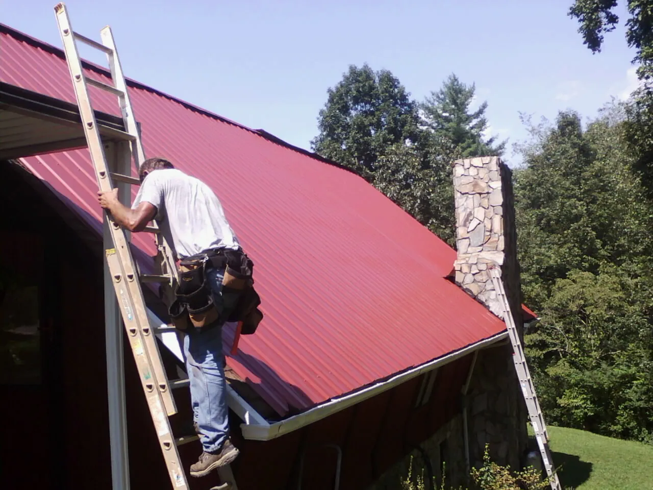 Roofer on a ladder beside a red metal roof and stone chimney — Soffit & Fascia Repair by Roofing Brothers in South Woodstock